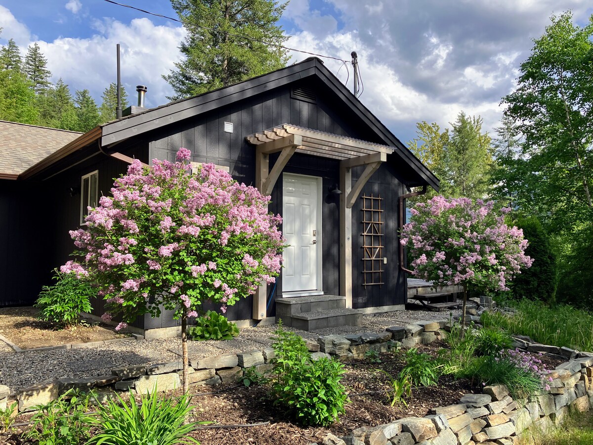 An exterior view of the B&B suite is presented, showcasing a dark-colored structure framed by lush greenery. Beautiful flowering trees are positioned symmetrically on either side of the entrance, leading to a welcoming white door. The surrounding landscape includes well-maintained garden beds and stone pathways.