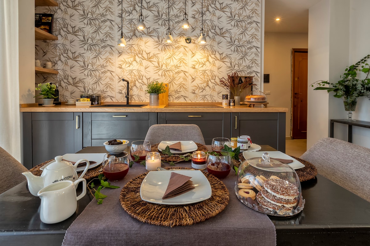 A dining table set for a meal features plates, cups, and candles, surrounded by natural decor. A well-organized kitchen with a wooden countertop and modern lighting is visible in the background, accented by greenery and patterned wallpaper.