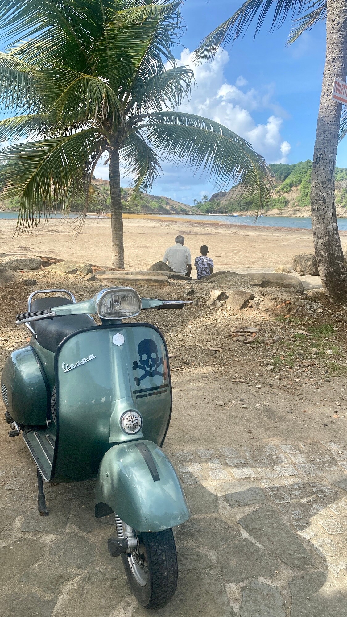 A vintage green scooter is positioned on a rocky path, with the beach and palm trees visible in the background. Two people are seated on a large rock, facing the calm waters of the bay, with hills and a partly cloudy sky framing the serene scene.