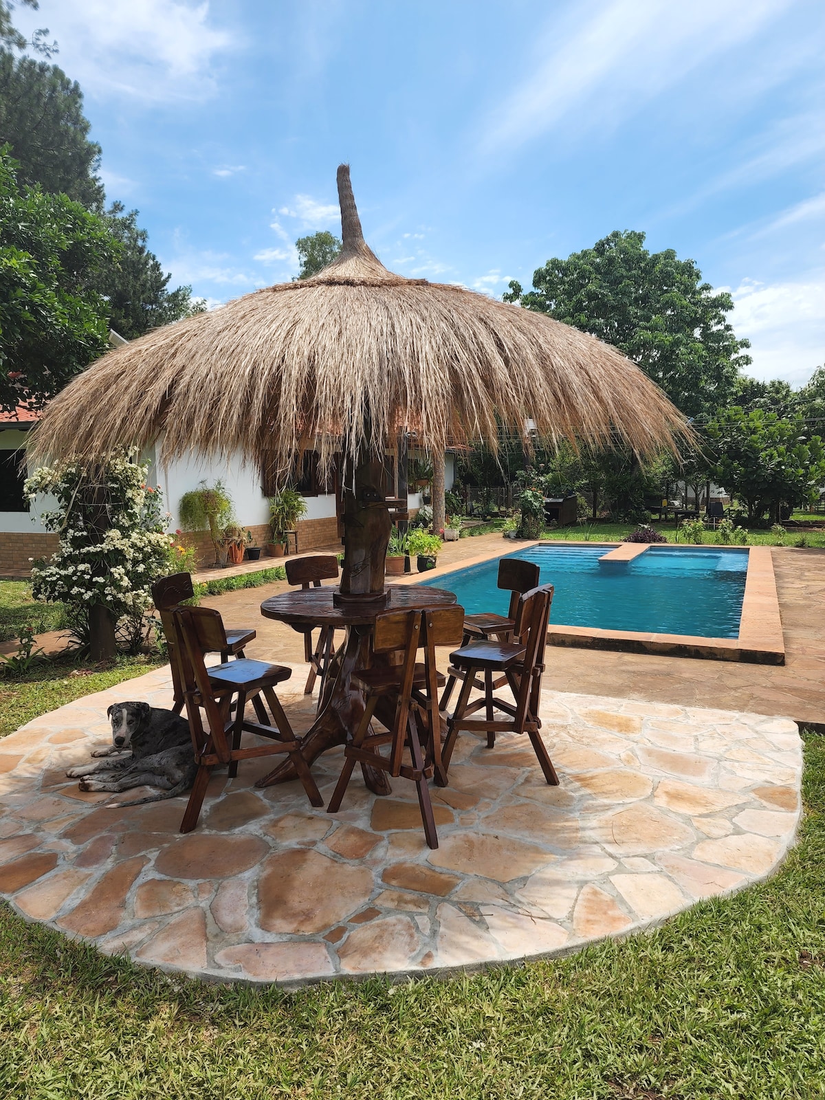 A thatched-roof gazebo is situated on a stone patio, accompanied by four wooden stools around a round table. The backdrop features a tranquil swimming pool surrounded by greenery, with a well-maintained lawn adding to the serene environment.