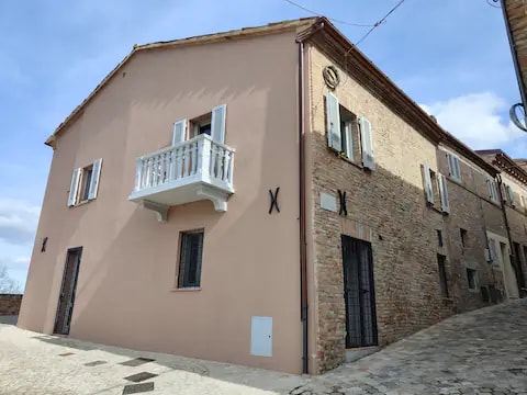 The exterior of the building showcases a blend of stone and stucco, featuring a distinct pastel pink façade. A small balcony with white railings is visible on the second floor, complemented by white shutters. Ground-level windows are adorned with black bars, adding to the structure's character.