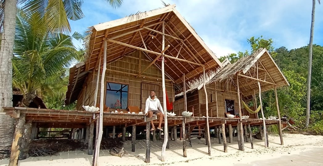 A traditional bamboo structure is set on stilts, featuring a spacious deck with seating. Large windows allow natural light to enter, and palm trees surround the home. A hammock adds to the relaxed ambiance, connecting the dwelling to the beachside environment.