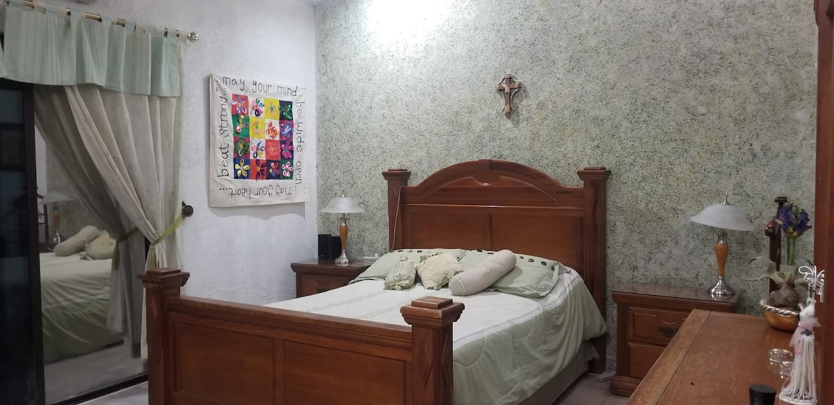A spacious bedroom features a large wooden bed with light bedding and decorative pillows. Two bedside lamps are placed on wooden nightstands. A decorative wall hanging adds color to the room, and a mirror reflects the gentle lighting from above.