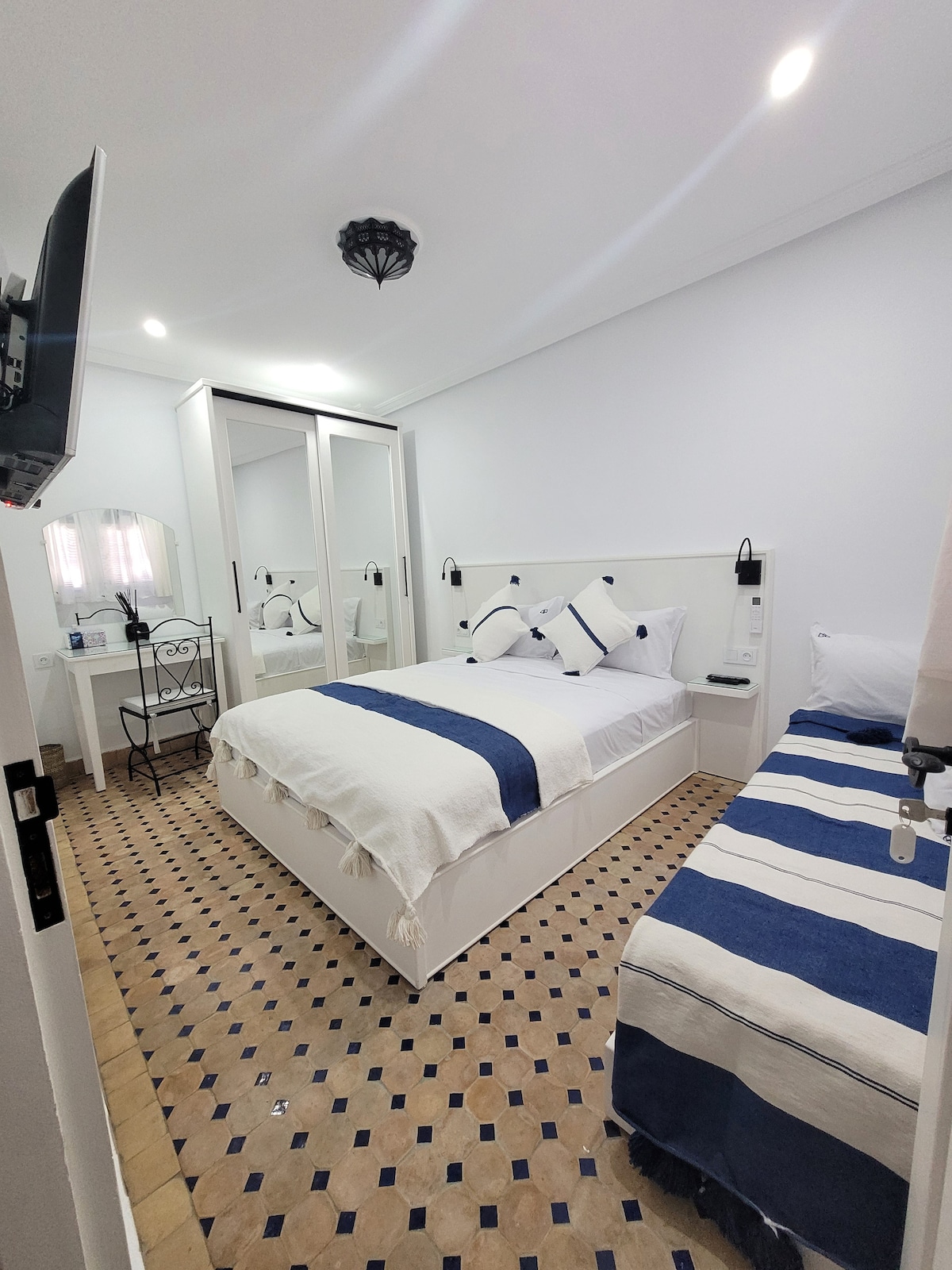 A well-lit bedroom features a spacious bed and an additional smaller bed, both adorned with navy and white striped bedding. A mirrored wardrobe reflects light, enhancing the room's brightness. A small table with a chair is positioned beside the window, adding functionality to the space.