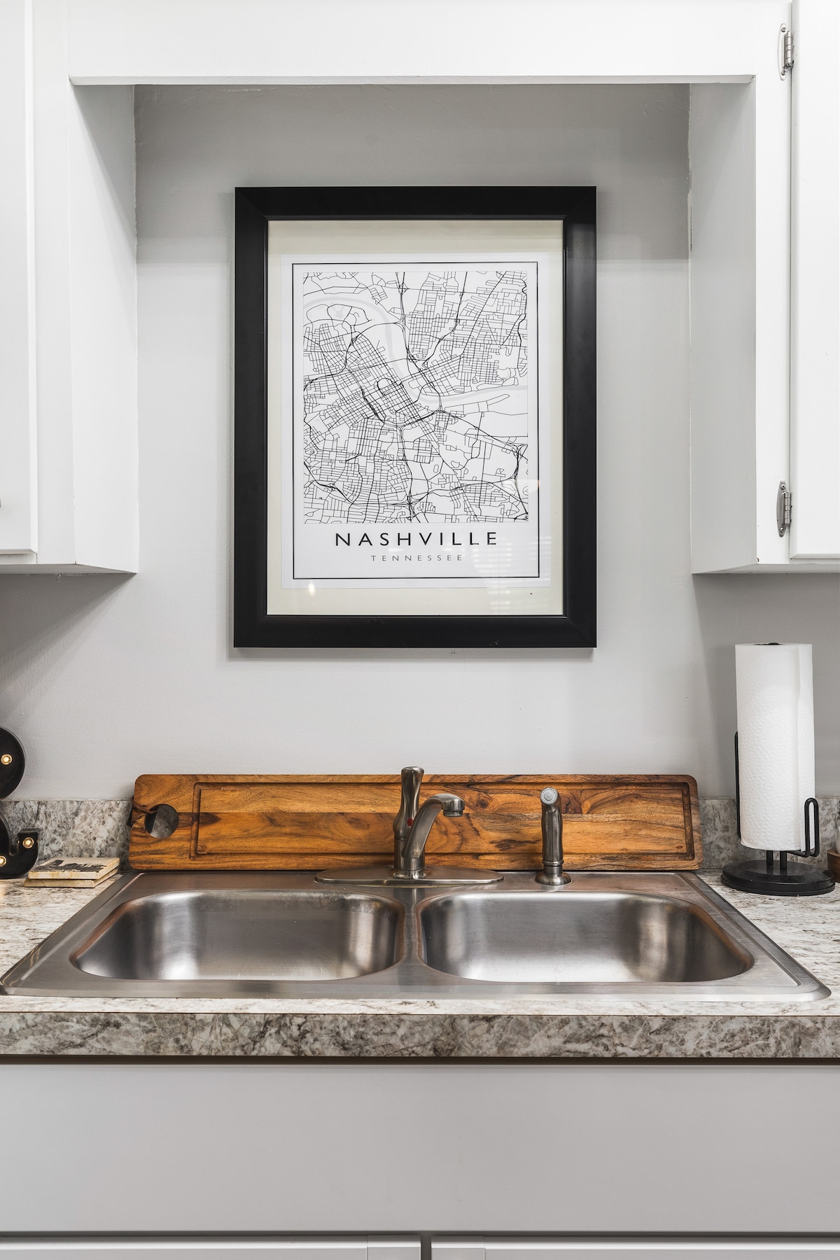A modern kitchen sink area features a double stainless steel sink with a brushed metal faucet. Above, a framed black-and-white map of Nashville is displayed on the wall, complemented by wood accents on the countertop.