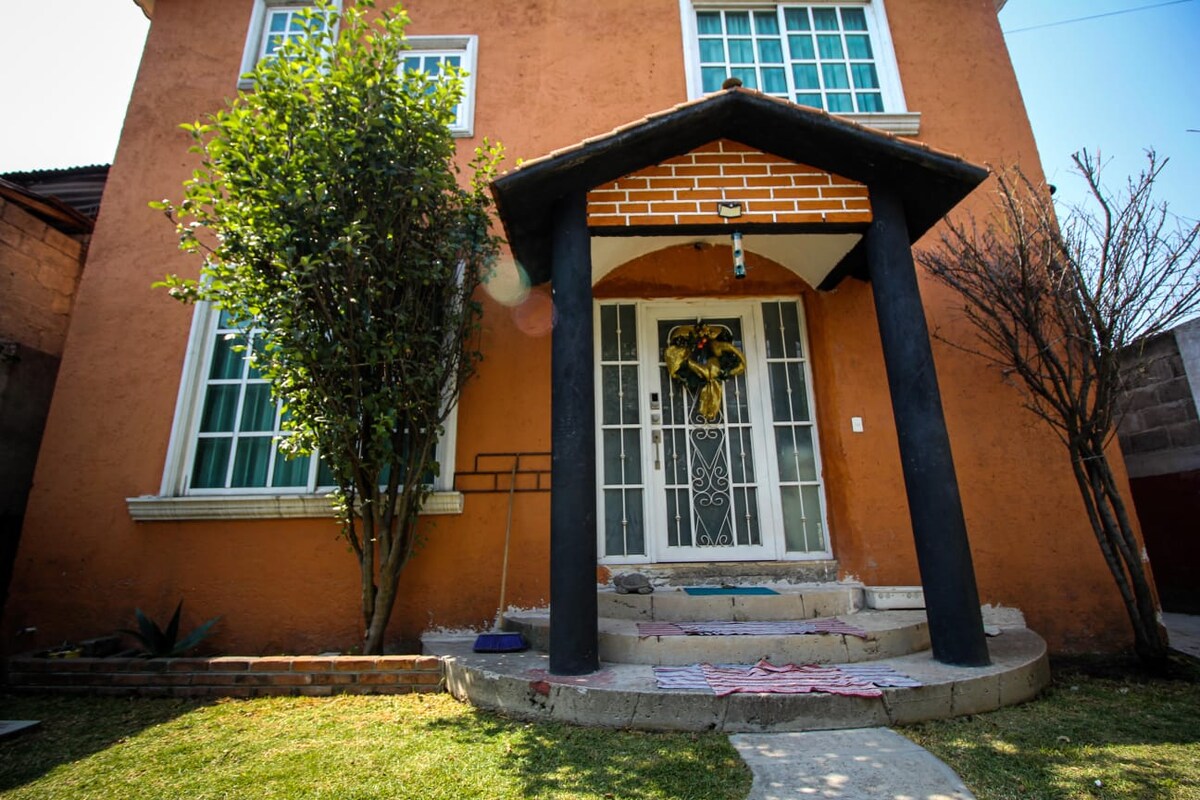 The exterior of a three-level house is visible, featuring an orange facade and a covered entrance. Large windows allow natural light, and a decorative wreath adorns the door. A well-maintained lawn is bordered by shrubs, enhancing the welcoming appearance of the home.