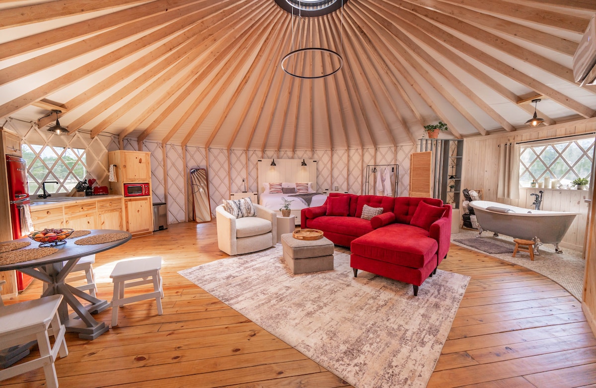 The spacious interior of the Yurt features high, wooden ceilings that enhance the open concept layout. A vibrant red plush sofa and matching armchair create a cozy seating area, complemented by a light-colored area rug. A beautifully designed Clawfoot tub is visible, adding character to the relaxing space.