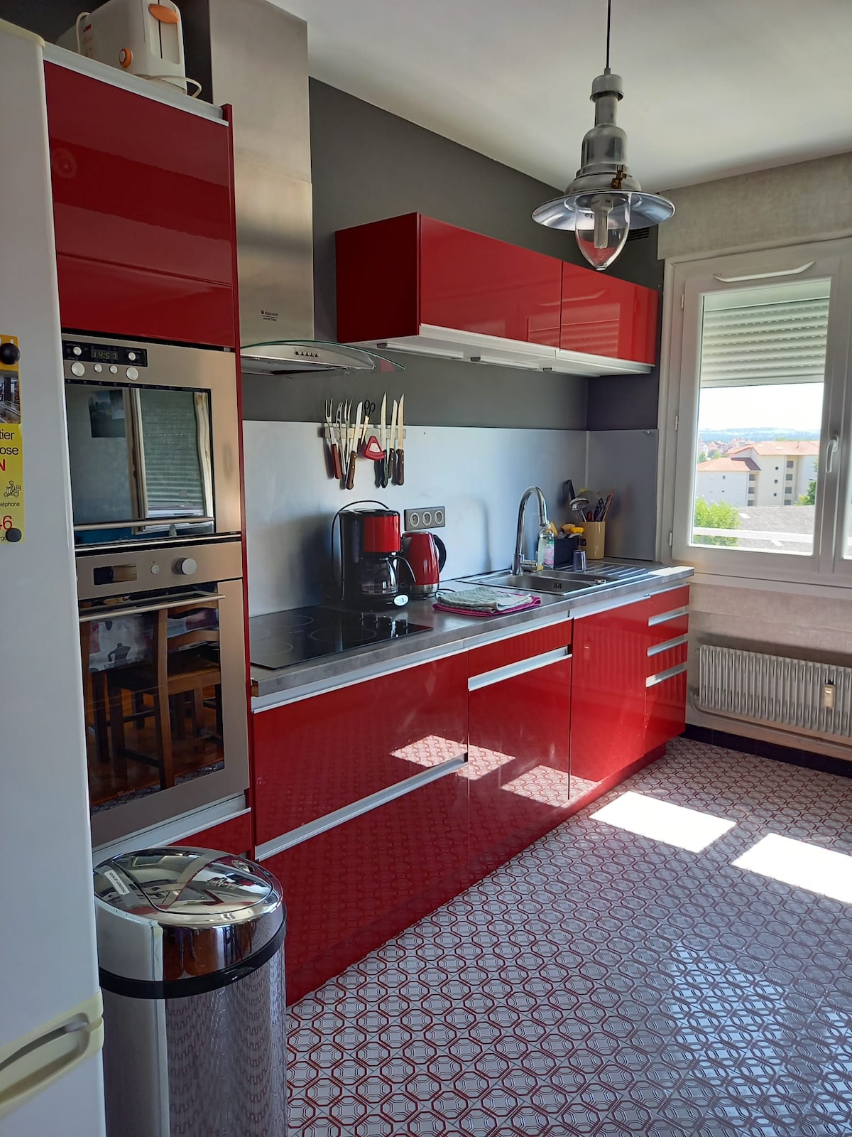 A modern kitchen features sleek red cabinetry and stainless steel appliances. Counter space is maximized with a combination of cabinets above and below. A small dining area with stools is visible, and sunlight brightens the space through a window, highlighting the patterned tile floor.