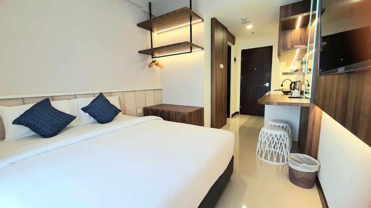 A well-lit bedroom features a spacious bed with crisp white linens and two contrasting blue decorative pillows. Minimalist wooden shelves and a compact kitchenette create an organized layout. The room is enhanced by smooth tile flooring and neutral-colored walls, contributing to a clean and modern aesthetic.