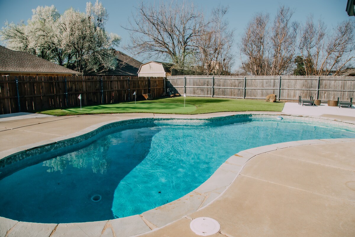 A spacious backyard features a swimming pool surrounded by a concrete deck. The pool is positioned next to a well-maintained putting green, with a wooden fence providing privacy. Leafless trees and residential homes are visible in the background, under a clear blue sky.