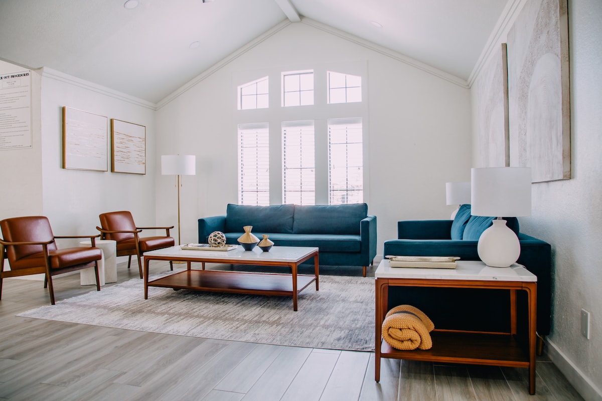 A spacious living room features two comfortable chairs and a sectional sofa, all arranged around a central coffee table. Large windows allow natural light to fill the space, enhancing the minimalistic decor, which includes a soft area rug and elegant side tables.