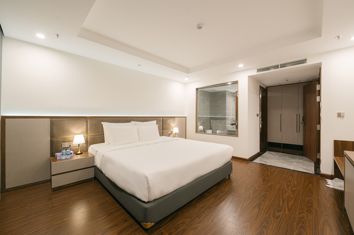 A spacious guest room features a large bed with crisp white linens centered between nightstands. A stylish bathroom area with glass partition is visible. Wooden flooring provides a warm feel, complemented by soft overhead lighting illuminating the neutral color palette.