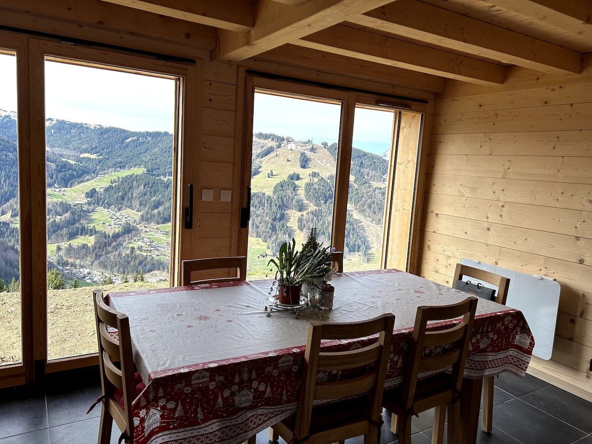 A dining area is seen with a wooden table surrounded by chairs. Large windows allow for an unobstructed view of the mountainous landscape. Natural light floods the space, and a small potted plant is placed in the center of the table, enhancing the atmosphere.