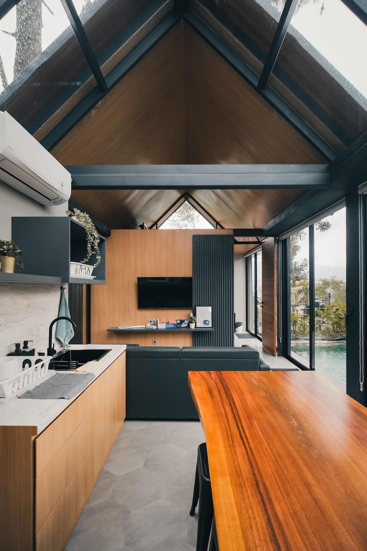 A modern kitchen area features sleek cabinetry and a minimalist design, complemented by a dining table made of rich wood. Large windows provide natural light and views of the surrounding nature, while a small living space is visible in the background, equipped with a wall-mounted TV.