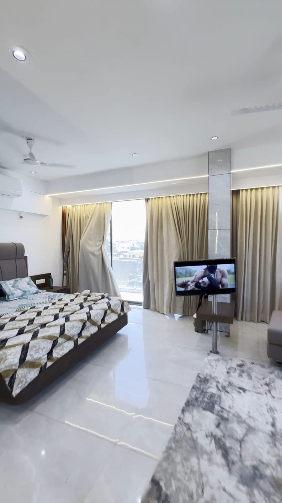 A modern bedroom features a spacious bed with a patterned quilt, accompanied by a flat-screen TV on a stand. Natural light enters through large windows, adorned with light curtains, creating a well-lit environment. A soft seating area is visible in the corner.