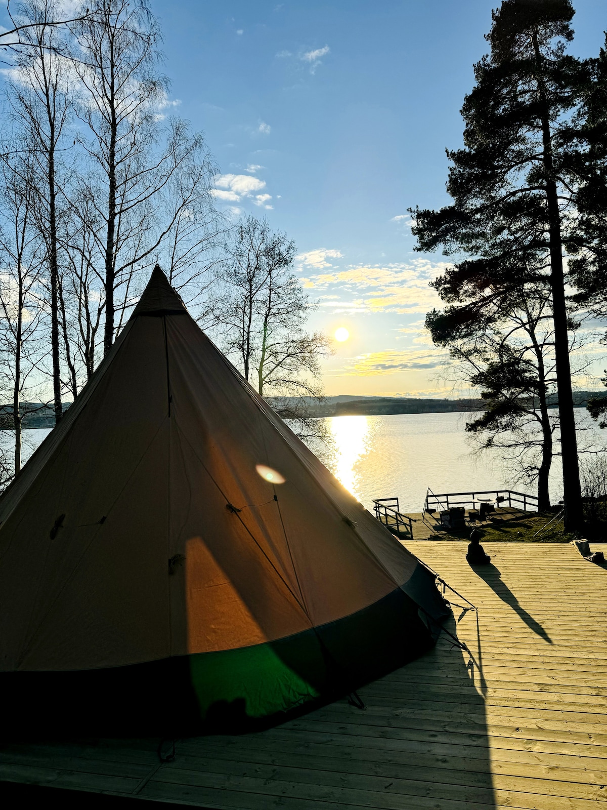 A spacious tent is situated near a calm lake, with sunlight reflecting on the water's surface. Trees frame the scene, and a wooden dock is visible in the background, enhancing the natural setting.