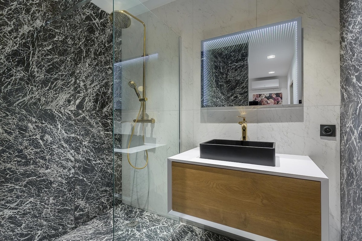 The modern bathroom features elegant marble walls in contrasting black and white. A sleek, minimalistic washbasin atop a wooden vanity is complemented by a stylish golden faucet. A walk-in shower with a glass enclosure showcases a minimalist design, offering a spacious and luxurious feel.
