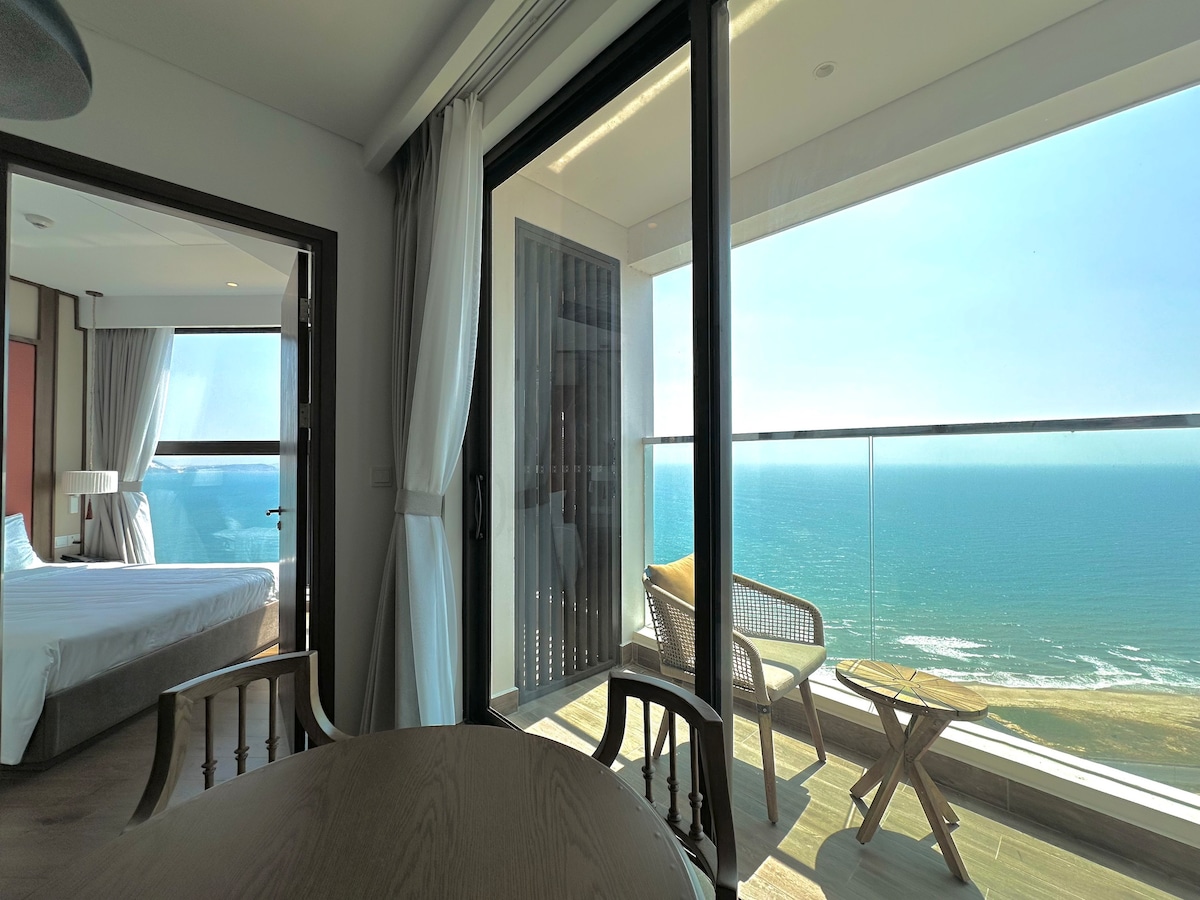 A bright living space features large glass doors that open to a balcony with ocean views. Comfortable seating is provided on the balcony, with sunlight illuminating the interior area, which includes a table and chairs, along with a spacious bed visible in the background.