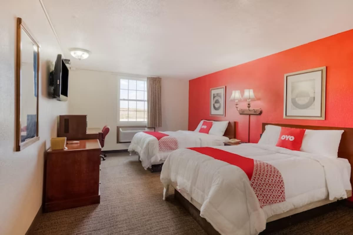 A well-lit room features two spacious beds dressed in crisp white linens. A vibrant red accent wall enhances the atmosphere. A large window allows natural light to fill the space, while a wooden desk and chair provide a dedicated workspace. A flat-screen TV is mounted on the wall.