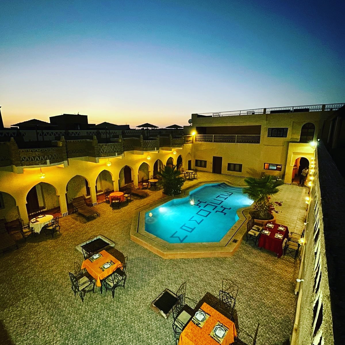 An expansive courtyard is highlighted by a swimming pool at its center, surrounded by arched doorways. Dining tables set with utensils are positioned around the pool, while warm lights illuminate the area, creating a relaxing evening ambiance.