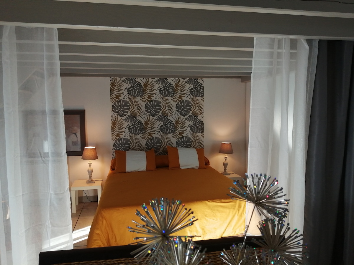 A cozy sleeping area features a spacious bed dressed in a vibrant orange coverlet, framed by light sheer curtains. Two bedside lamps illuminate the space, contrasting with a decorative headboard adorned with a leaf pattern.
