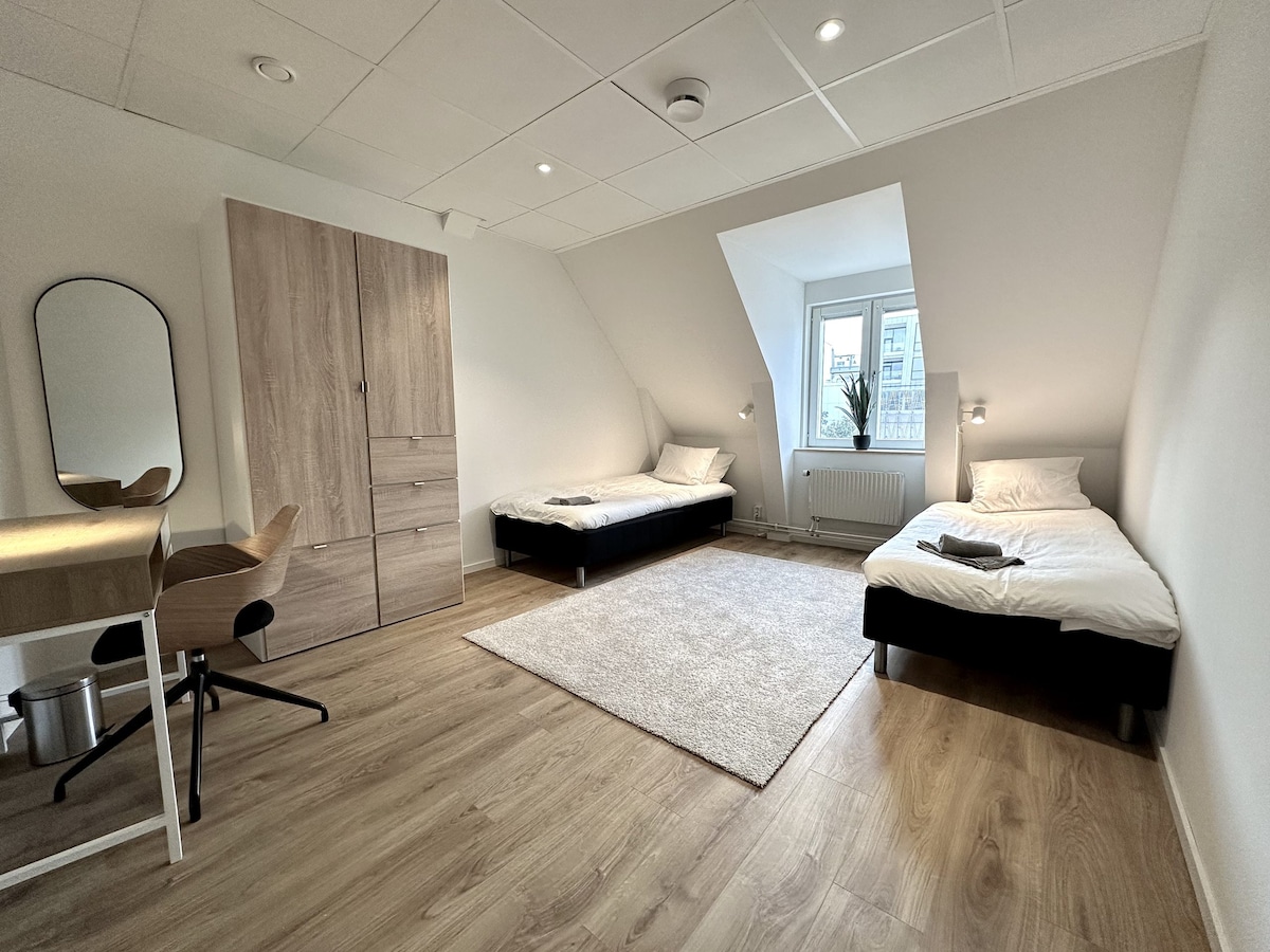 A bright room features two single beds positioned beside a soft area rug. A desk with a chair is placed against one wall, complemented by a mirror. A window offers natural light, while a wardrobe provides storage space. Light wood flooring adds warmth to the setting.