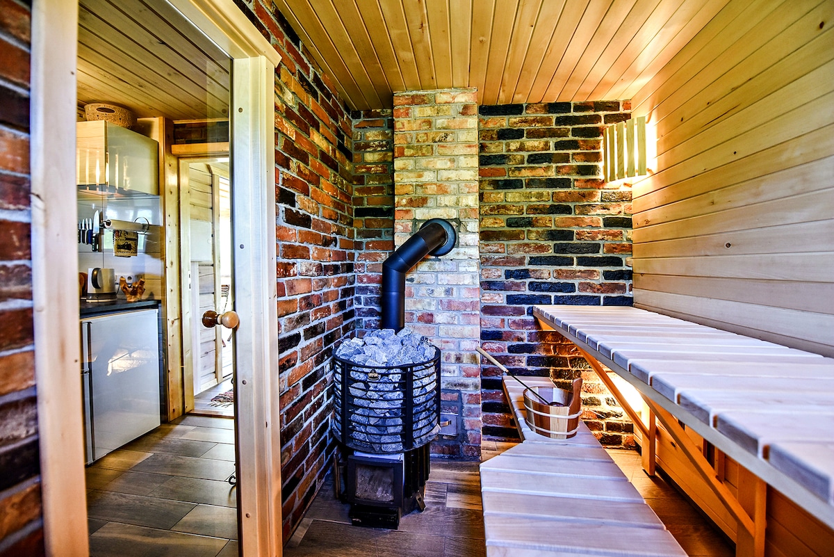 A rustic interior reveals a brick wall feature, complementing the warm wooden finishes. A wood-burning stove, adorned with stones, is positioned beside a wooden bench. Soft lighting from wall sconces enhances the cozy atmosphere. The entrance to a kitchen area is visible in the background.