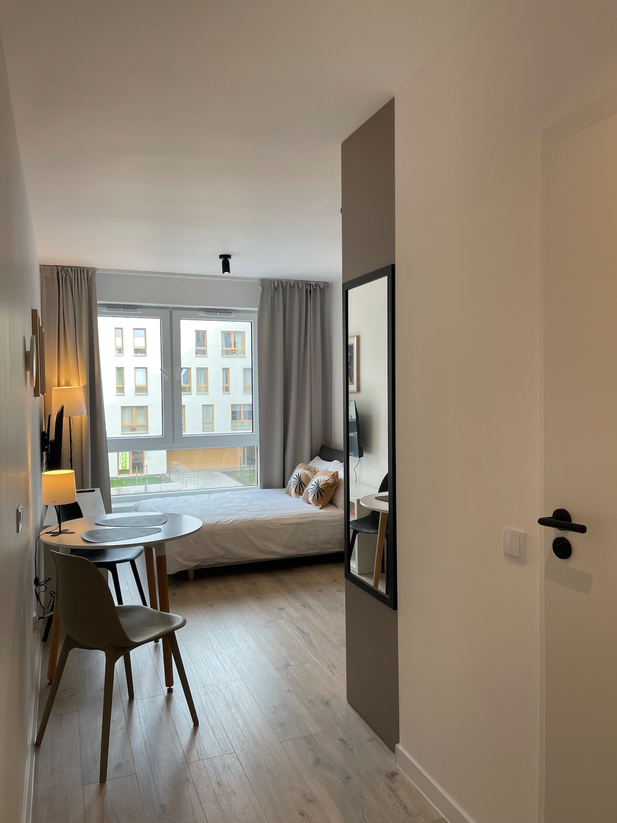 A well-lit bedroom is visible through a doorway, featuring a spacious bed with decorative pillows. A small round table and a chair are positioned across from the bed. Natural light enters through a large window, complemented by light-colored curtains.
