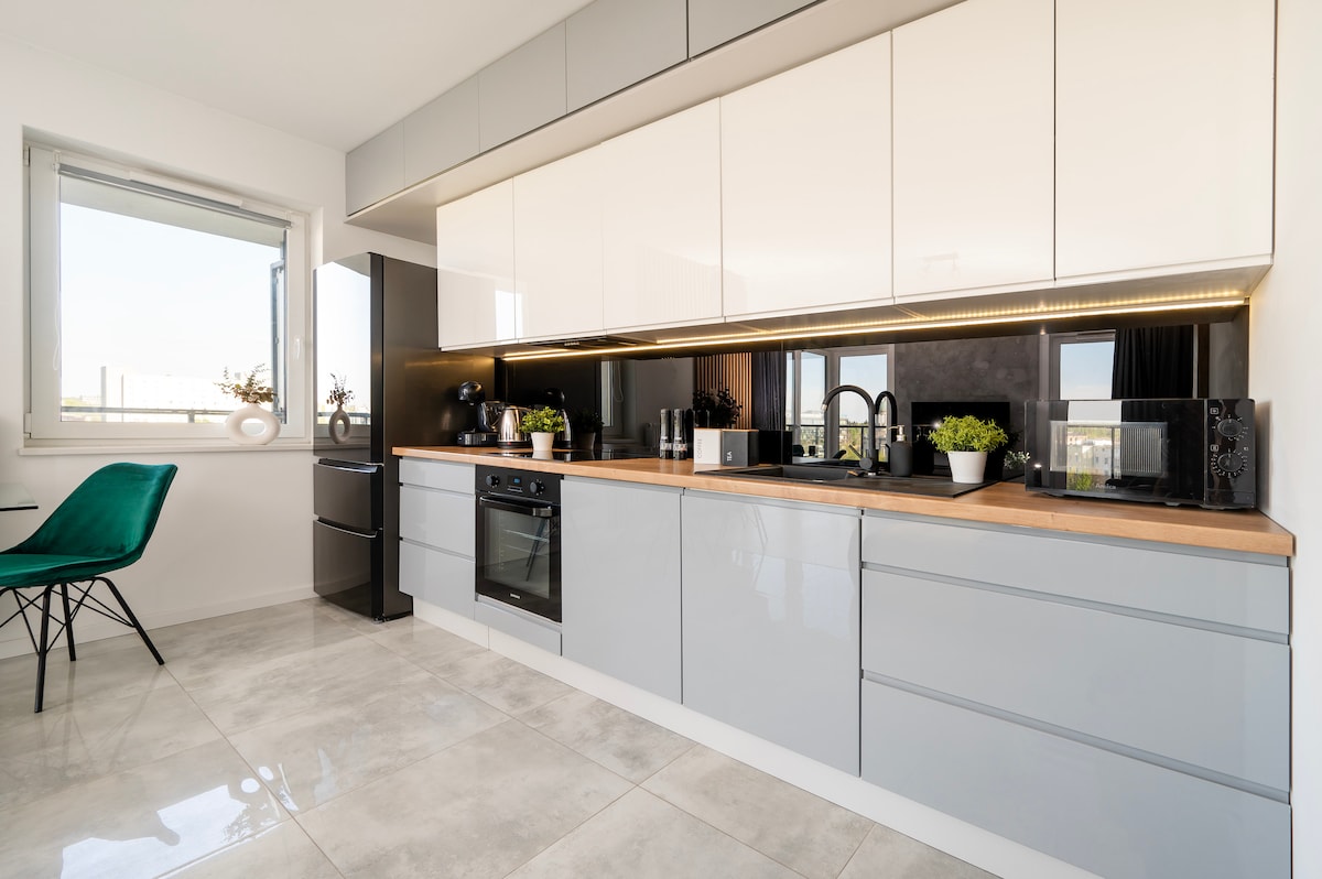 A modern kitchen is presented with sleek cabinetry in gray and white, featuring a wooden countertop. Appliances include an oven, stovetop, and refrigerator. Natural light enters through a window, highlighting the minimalist design. Potted plants add a touch of greenery to the space.