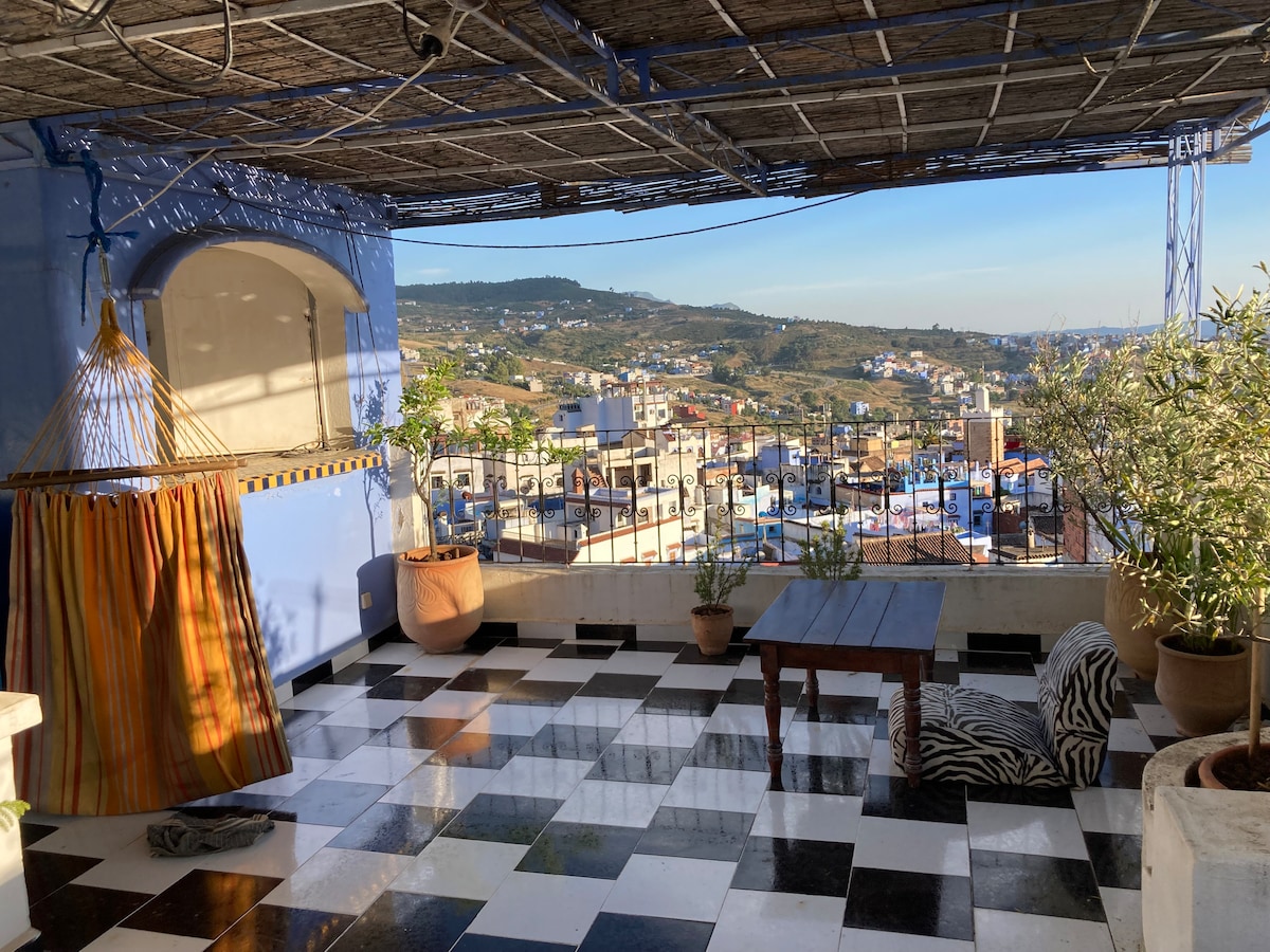 A spacious terrace with a checkered floor design is shown, featuring a comfortable hammock draped in colorful fabric. Potted plants add greenery to the area, while views of the surrounding hillside and buildings can be seen against a clear sky.
