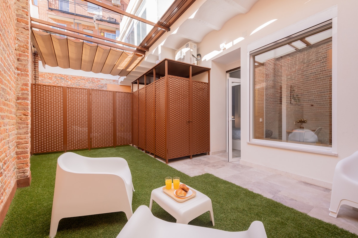 A private patio area features artificial grass and modern white seating. The space is enclosed by patterned wooden screens and has a glass door leading into the interior. Natural light filters in the open area, creating a serene outdoor environment.