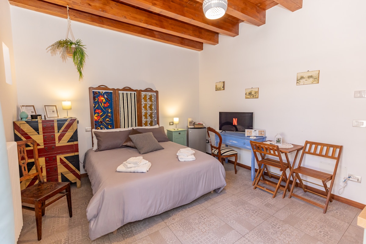 The cozy bedroom features a spacious bed with soft linens and decorative pillows. A small TV is positioned on a light blue desk, which is accompanied by two folding chairs. Warm wooden beams adorn the ceiling, adding a touch of charm.