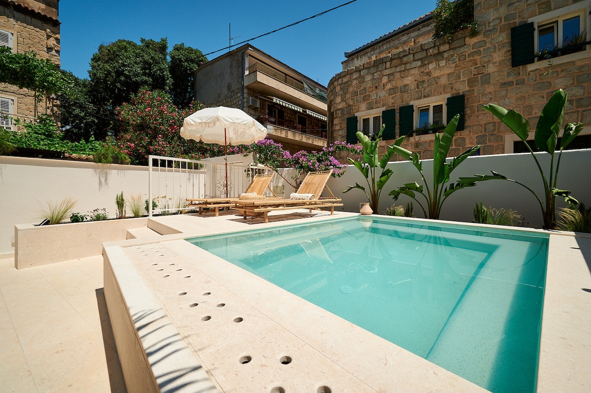 A shared outdoor space features a clear jacuzzi surrounded by sun loungers and a large umbrella. Lush greenery and flowering plants provide a natural backdrop, while stone walls and vibrant architectural details add character to the area.