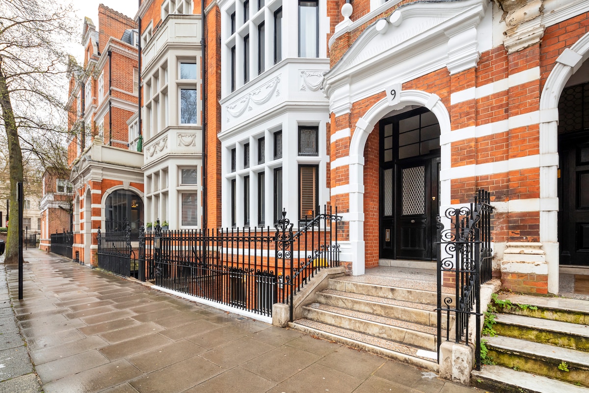 The exterior of a charming red-brick building is visible, featuring intricate architectural details and large windows. The entrance is highlighted by a set of stairs leading up to the front door, with a wrought-iron railing enclosing the property. A clean, paved walkway is seen in front.