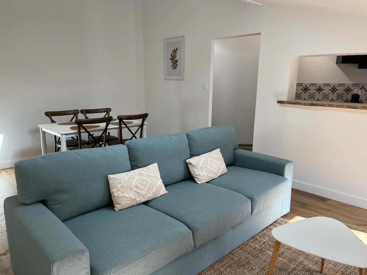 A modern living space is captured, featuring a comfortable sofa adorned with decorative cushions. In the background, a dining area with a table and chairs stands beside an open kitchen, characterized by a neutral colour palette and minimalistic decor.