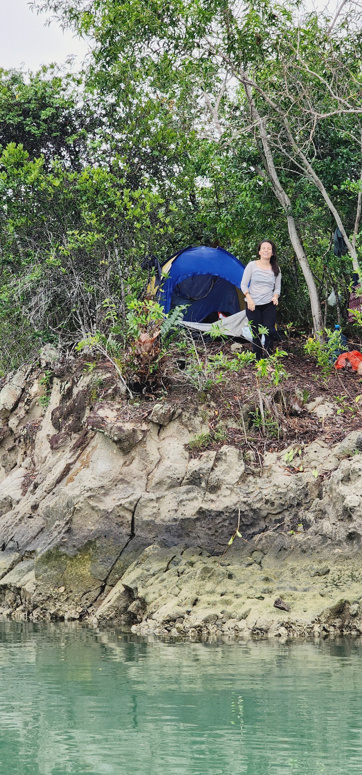 A bright blue tent is nestled among lush greenery on a rocky outcrop. The calm water reflects the surrounding landscape, enhancing the natural setting. A person is seen standing nearby, engaged in the tranquil environment, with trees framing the campsite.
