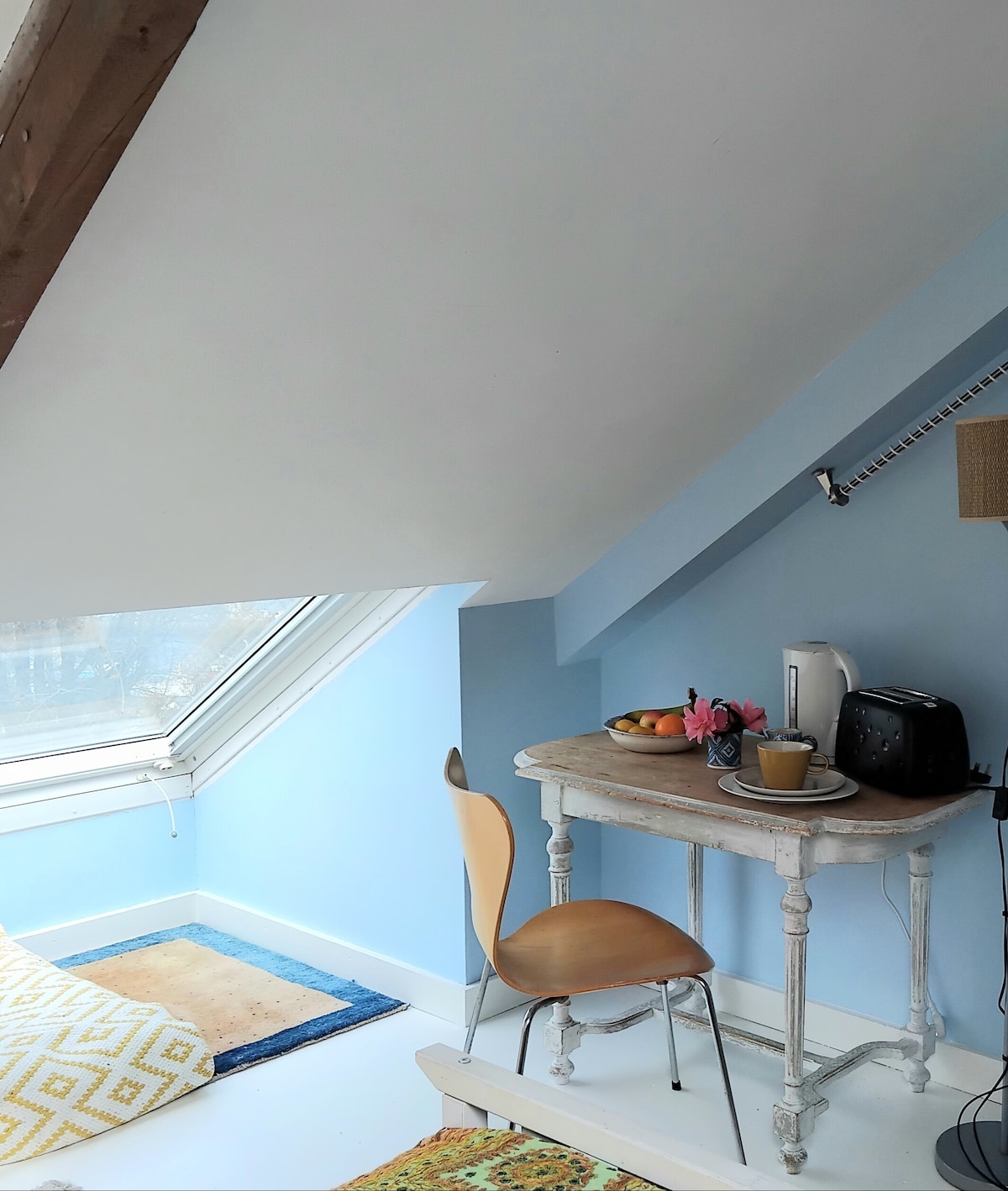 A cozy nook features a small table with a kettle and a coffee maker, accompanied by a chair. A vase with flowers enhances the space. Natural light filters through a large skylight, illuminating the blue walls and the patterned rug beneath the table.