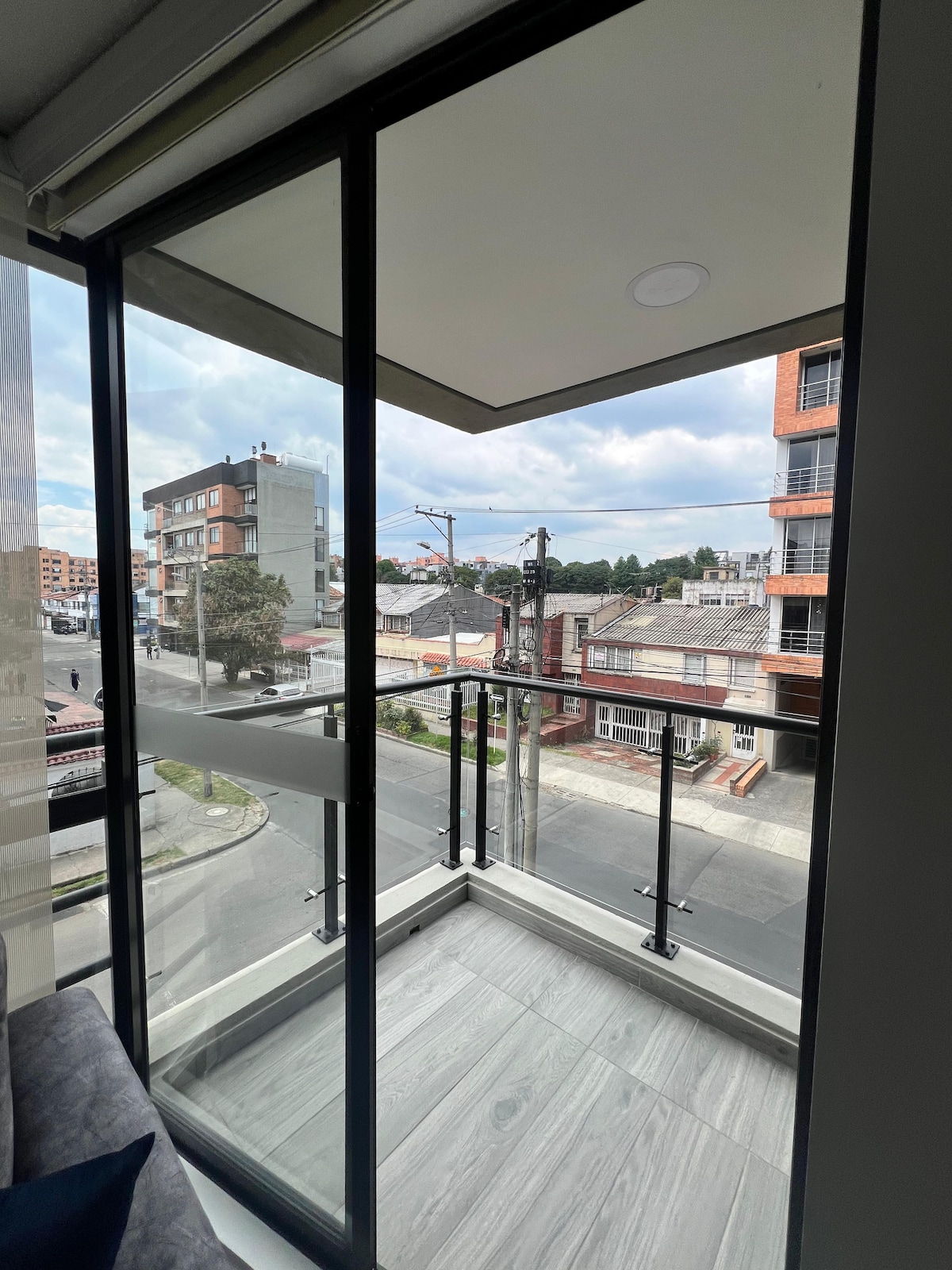 A balcony view presents a glimpse of the surrounding neighborhood, featuring a mix of residential buildings and tree-lined streets. Floor-to-ceiling glass doors enhance natural light, while the modern railing provides safety and openness to the outdoor space.
