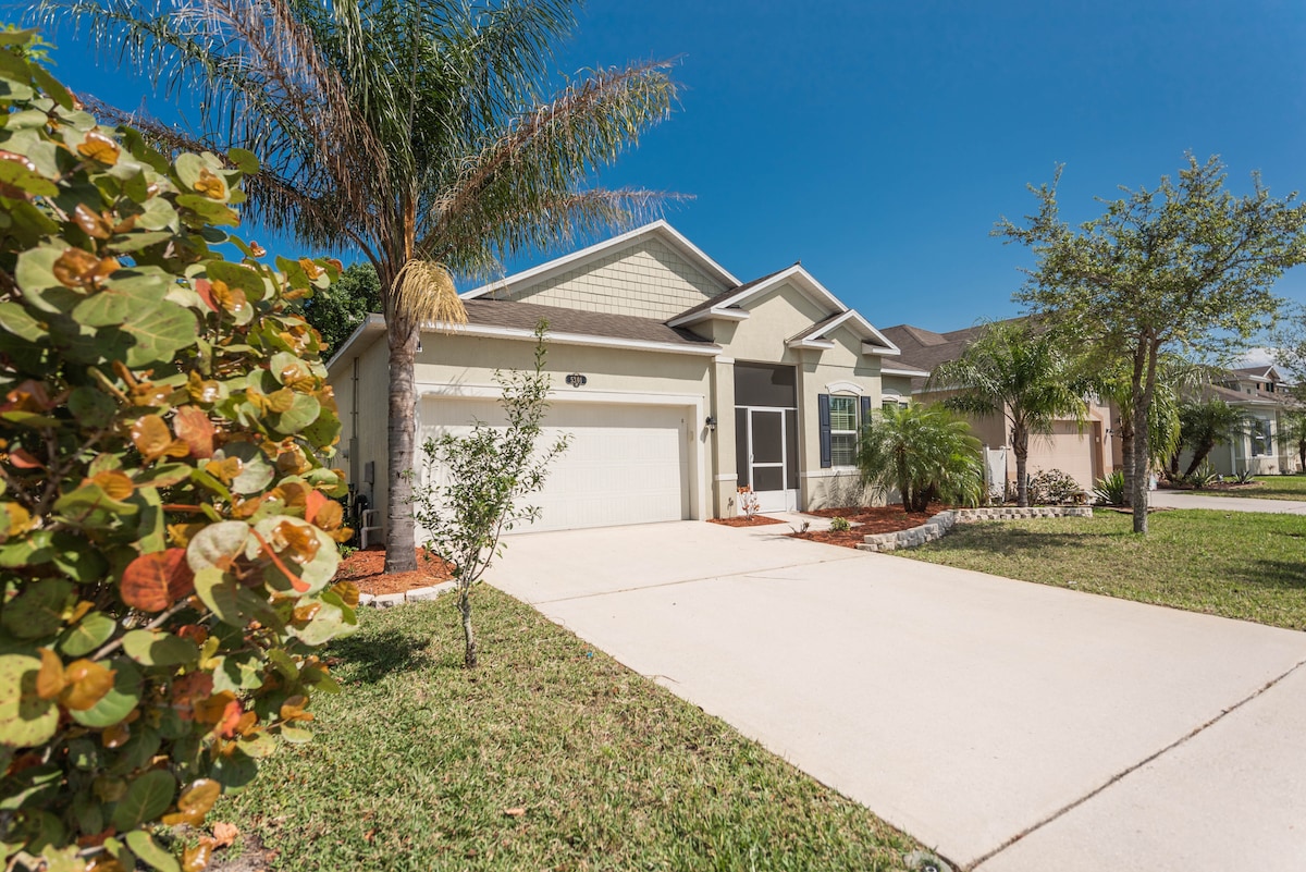 A single-story home is presented with a well-maintained front yard featuring lush green grass and shrubbery. A paved driveway leads to a two-car garage. Palm trees and other greenery are visible, offering a serene environment under a clear blue sky.
