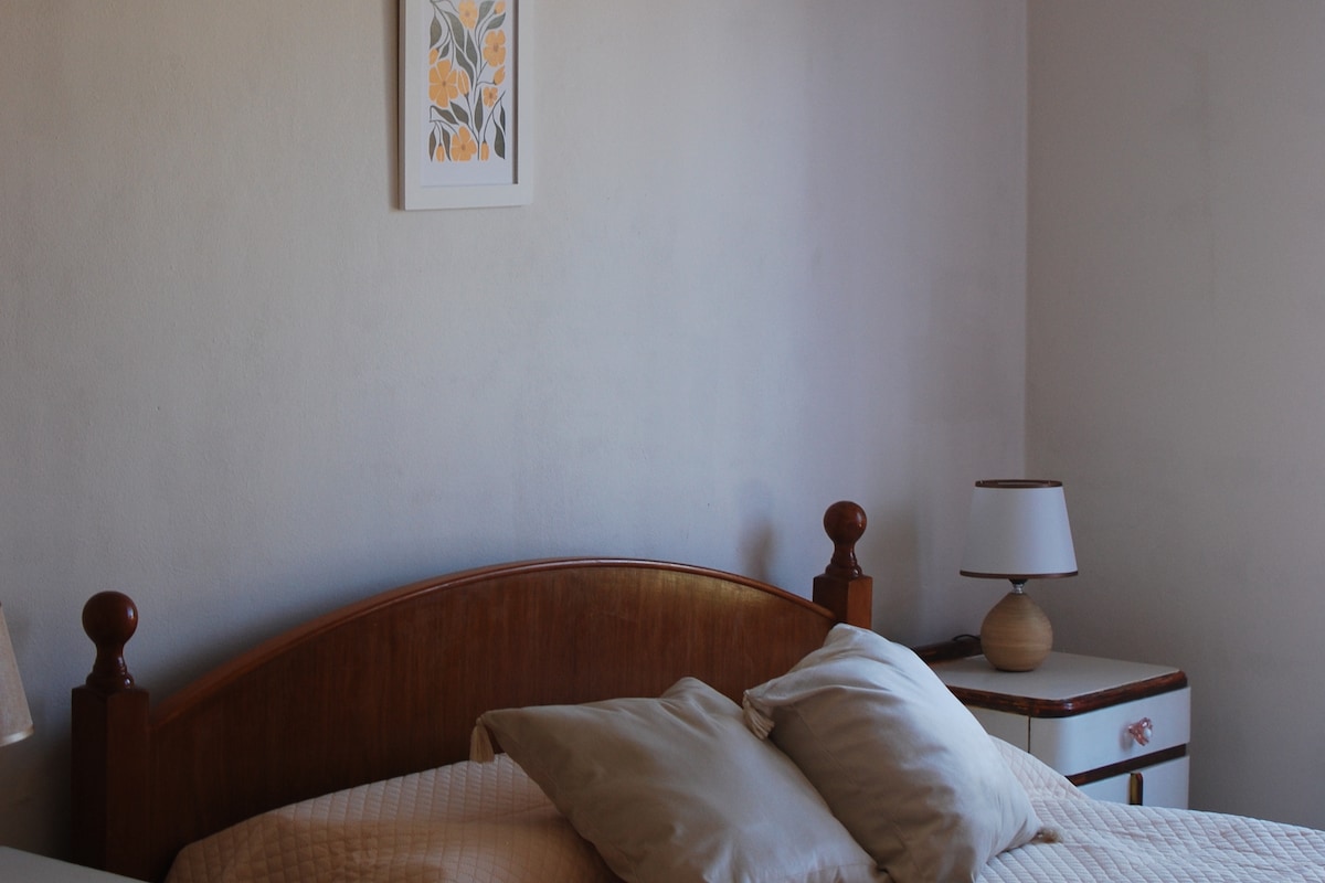 A cozy sleeping area features a wooden headboard with soft pillows arranged neatly. A bedside lamp stands on a small nightstand next to the bed, while a framed floral artwork adorns the wall above, contributing to a serene environment.