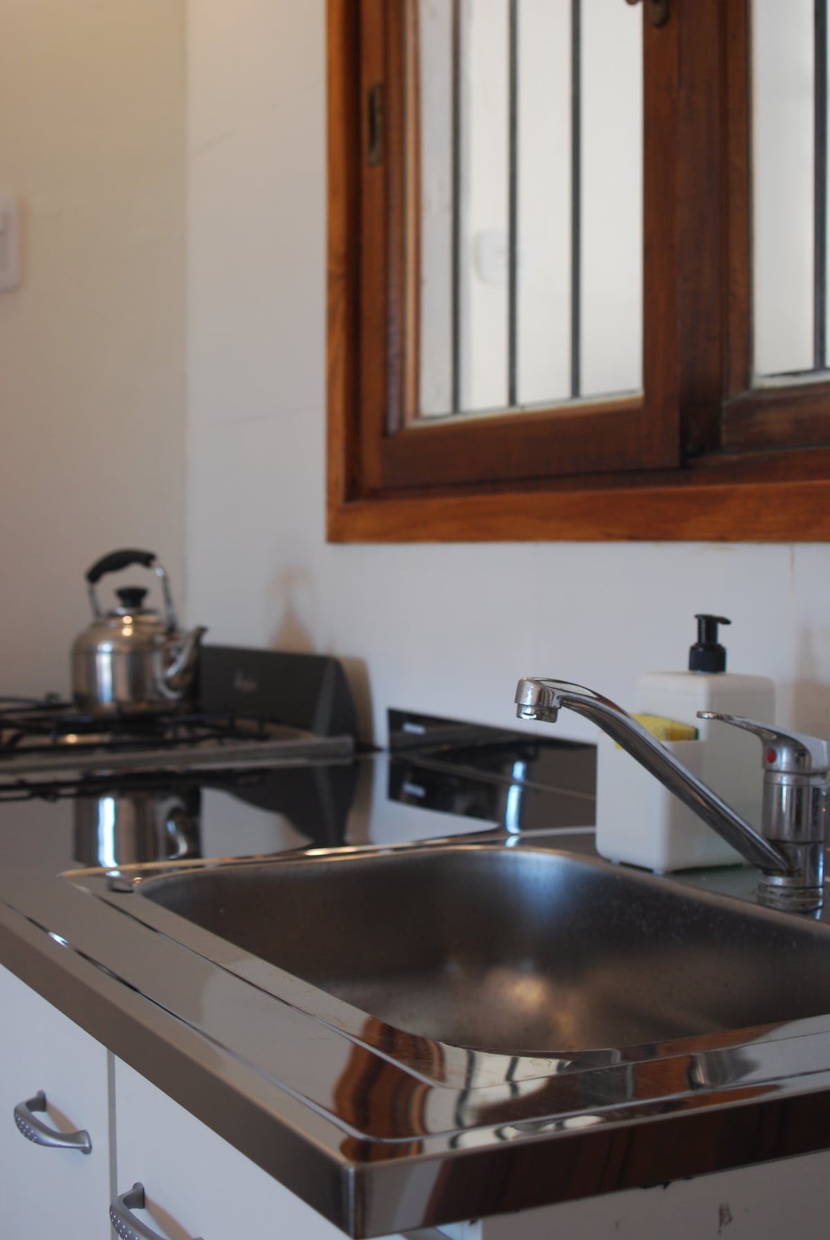 A compact kitchen area is visible, featuring a stainless steel sink and polished countertop. A kettle sits beside the stovetop, with a sleek gas burner offering a modern touch. Natural light filters in through a window with protective bars, enhancing the functional workspace.