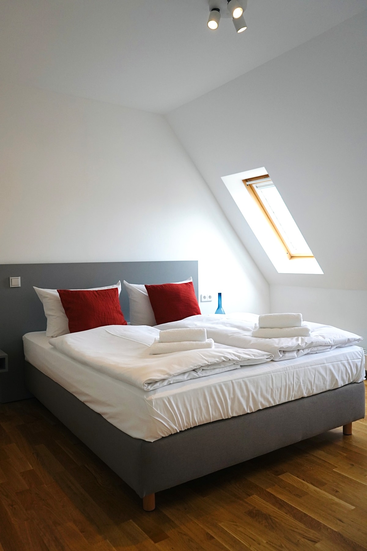 A cozy bedroom features a spacious bed adorned with white linens and two red accent pillows. Natural light enters through a slanted window, highlighting the wood flooring and minimalist design of the room.