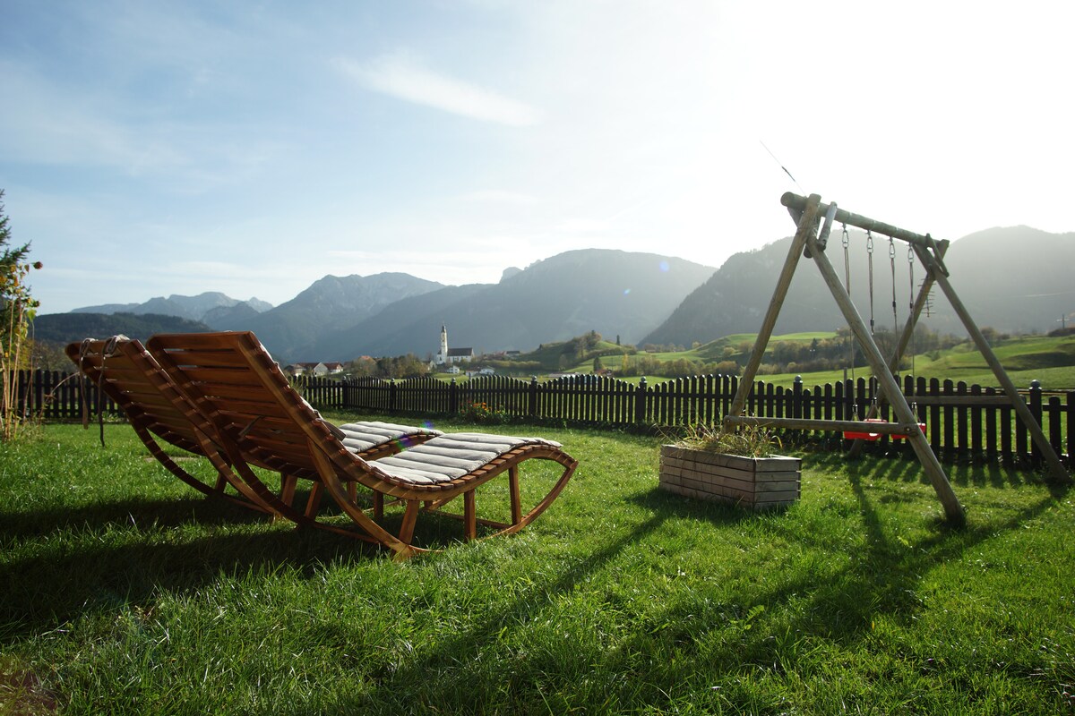Two wooden lounge chairs are positioned on a grassy lawn, facing expansive views of distant mountains and a clear sky. A simple swing set stands nearby, adding an element of playfulness to the outdoor area.
