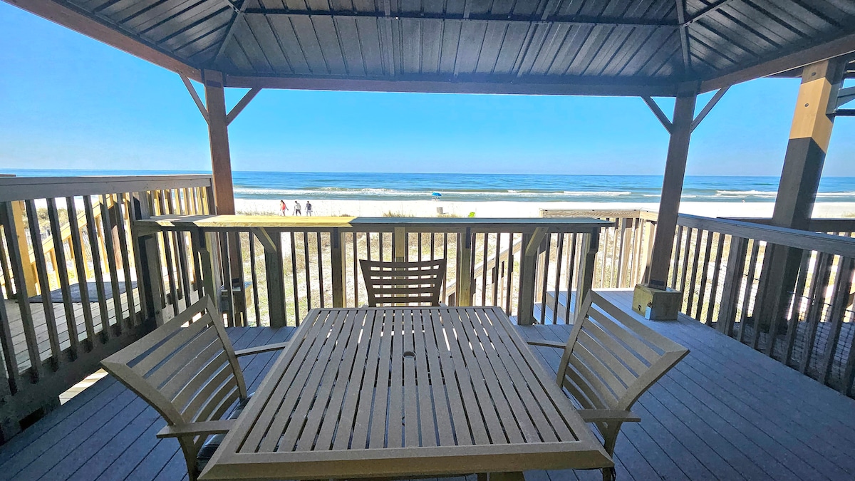 A covered deck area features a wooden dining table surrounded by several wooden chairs, all positioned with a clear view of the beach. The ocean waves are visible in the distance under a bright blue sky, creating a serene atmosphere for outdoor enjoyment.