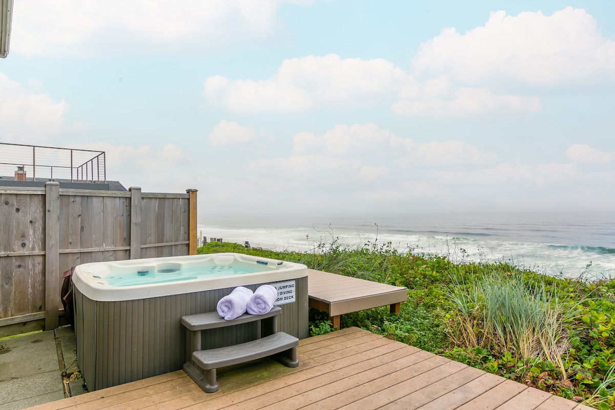 An oceanfront hot tub is situated on a wooden deck, with plush towels neatly arranged on its edge. Lush greenery surrounds the area, with a view of the ocean and rolling waves visible in the background under a soft cloudy sky.