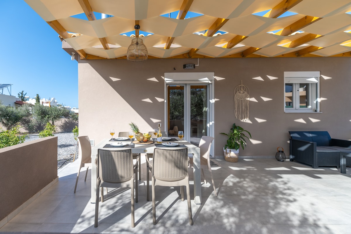 An outdoor dining area is comfortably furnished with a table and chairs under a lattice-patterned roof. The space is well-lit by natural light, and features plants and decorative elements, creating a relaxed and inviting atmosphere for al fresco meals.
