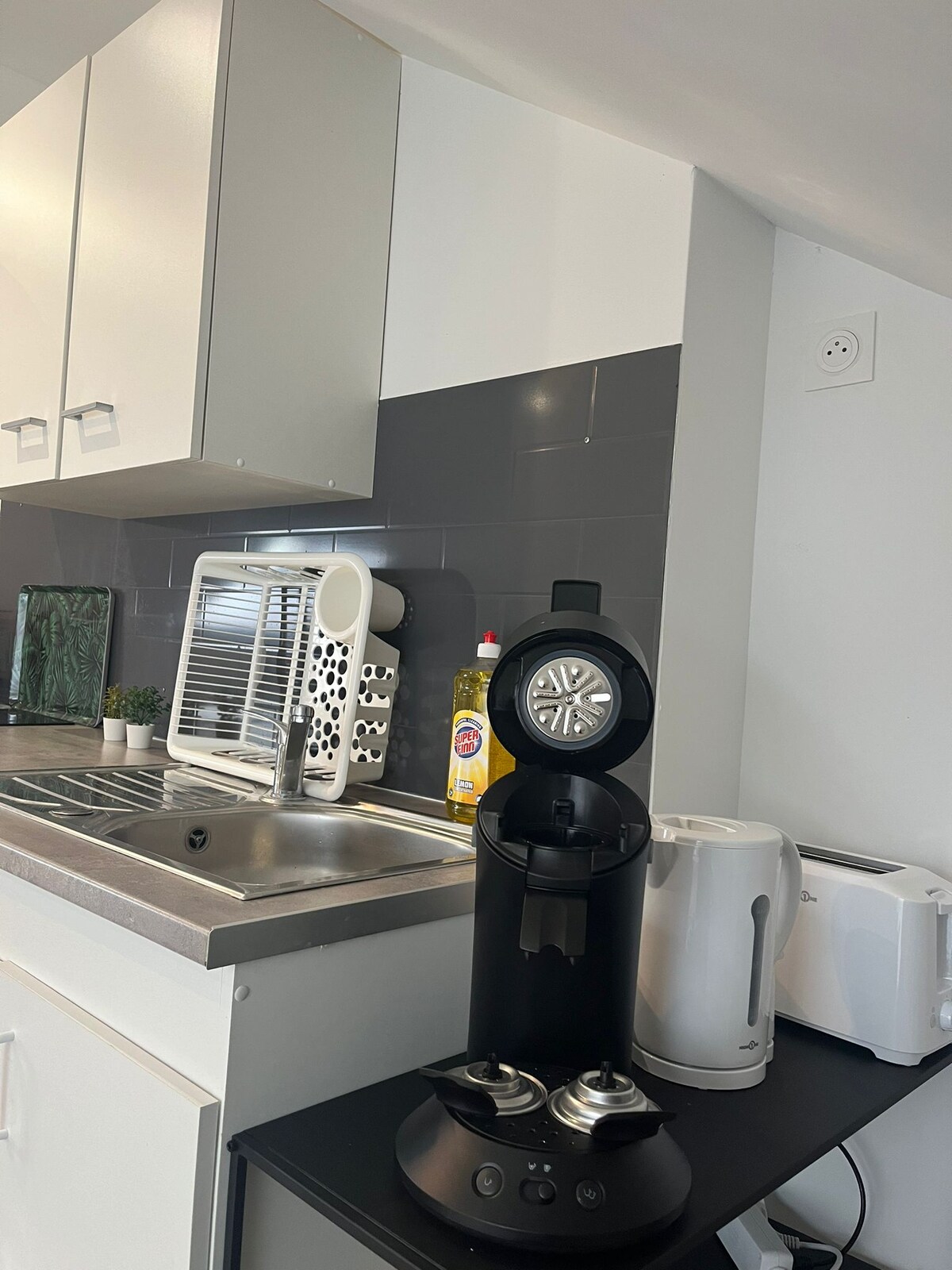 A modern kitchenette is presented, featuring sleek cabinetry and a stainless-steel sink. Essential appliances include a black coffee machine, an electric kettle, and a toaster, arranged neatly on a countertop. Natural light highlights the functional design and ample workspace.