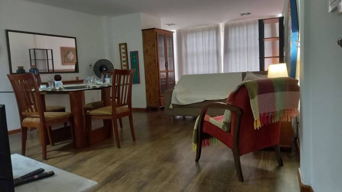 A spacious studio features a dining table set for four, complemented by wooden chairs. A comfortable armchair with a colorful throw is positioned nearby. Light filters through the sheer curtains, illuminating the room's minimalist decor, which includes a fan and wooden cabinetry.