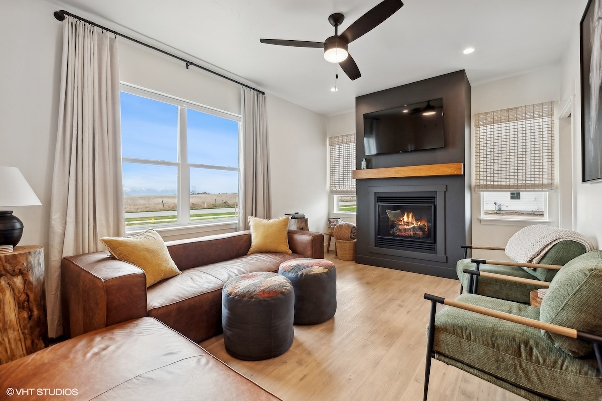 A welcoming living room features a sleek fireplace and a mounted television above. Comfortable leather and fabric seating options, including a sectional with rounded cushions, invite relaxation. Large windows provide natural light and views of the outdoors, while wooden accents add a modern touch.