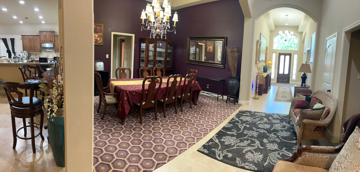 A spacious dining area is highlighted by a long, elegant table surrounded by dark wooden chairs. Rich purple walls provide a warm backdrop, while a chandelier adds soft lighting. The adjoining living area features patterned rugs and a stylish sofa, creating a cohesive and inviting space.